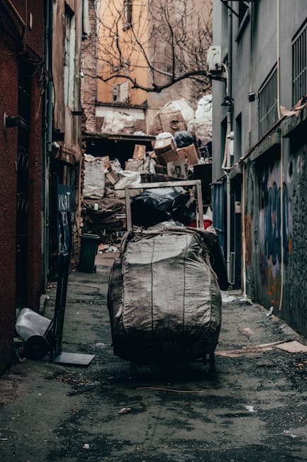 A narrow urban alleyway cluttered with discarded rubbish and debris, featuring a large, worn black trash bag in the foreground, which appears to be full and slightly open, revealing some contents. Beh