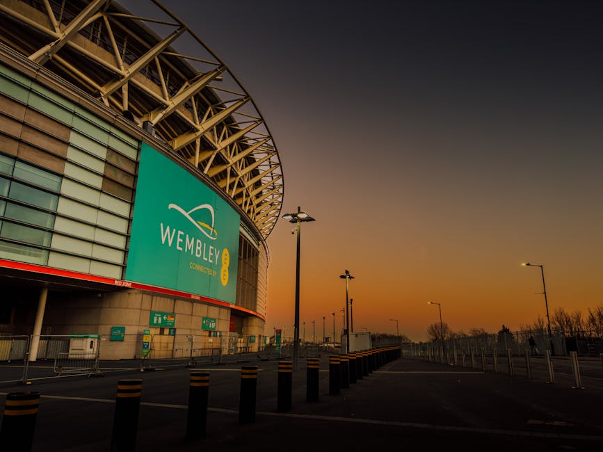 Inside Wembley Stadium during a football match, the image captures a wide view of the packed seating areas filled with spectators wearing various colours, predominantly red, black, and white. The lush